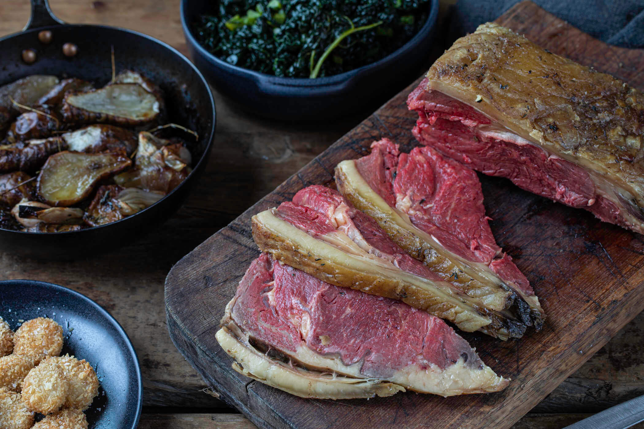 Bone-in sirloin roast sliced on a wooden board with Jerusalem artichokes, shallots, cavolo nero and crispy bone marrow balls