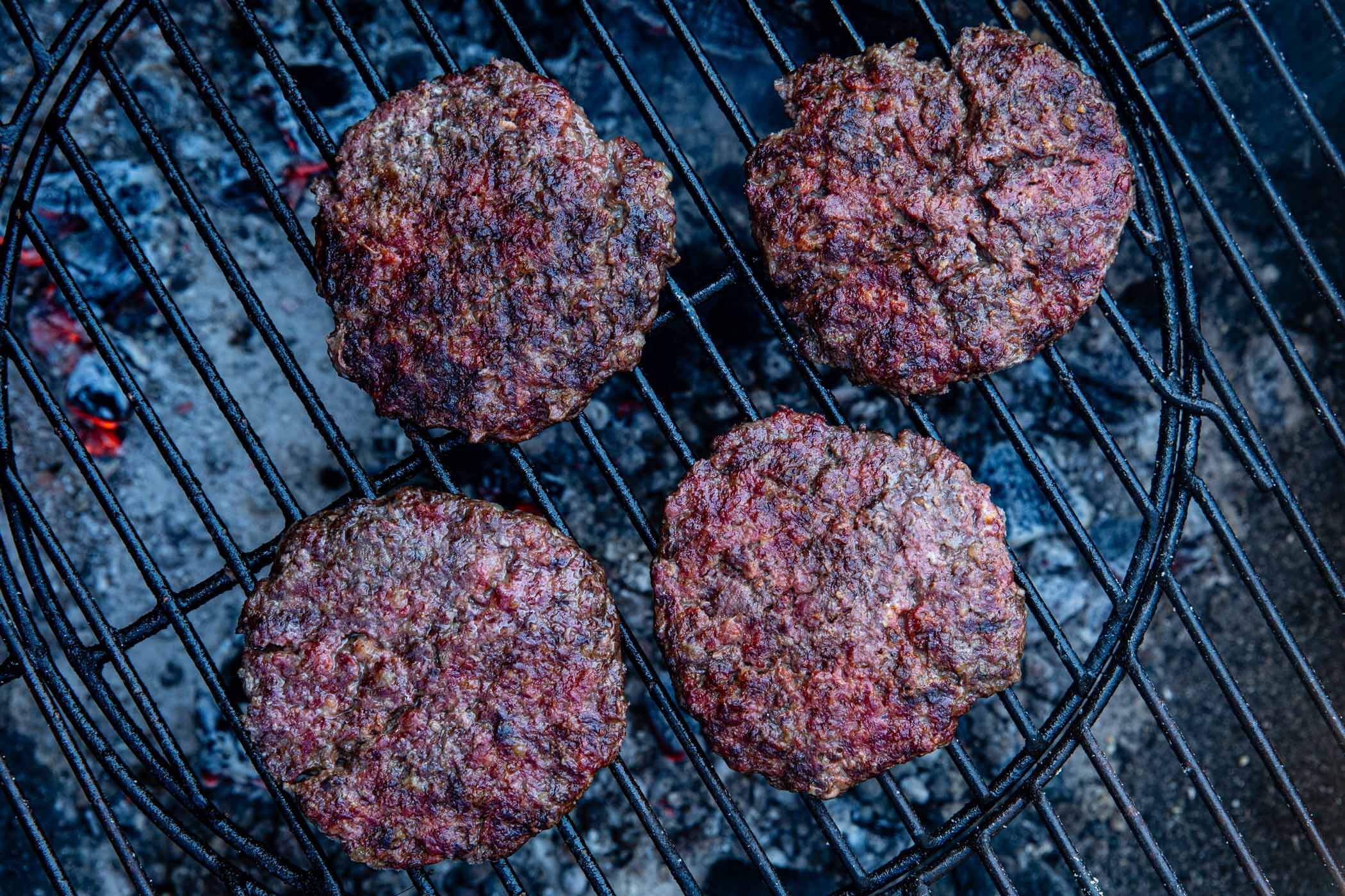 Four steak burgers cooking on a barbecue grill, with visible grill marks and a smoky char