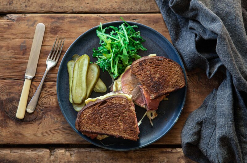 Corned beef ox tongue reuben sandwich cut in half, served with sliced gherkins and dressed rocket.