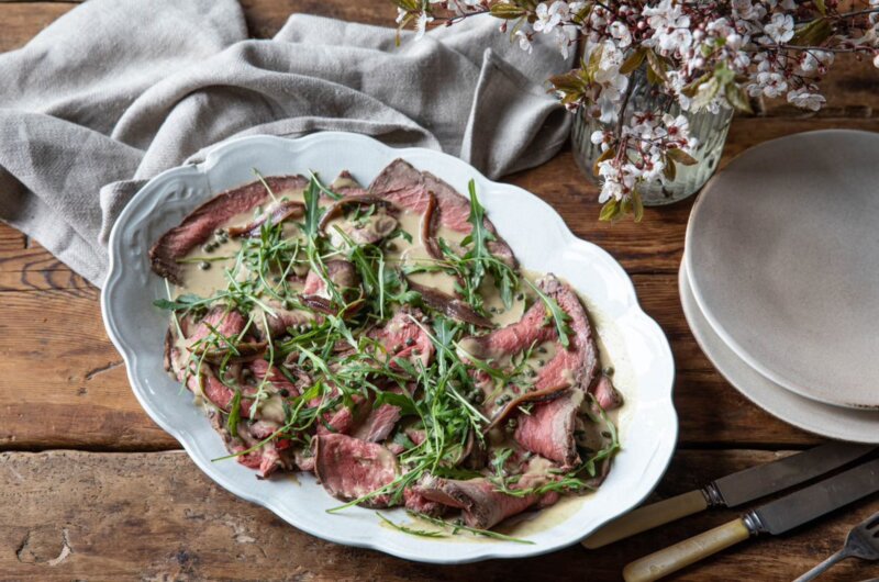 Close-up of rare roast topside of beef with tonnato dressing served in a white serving dish.