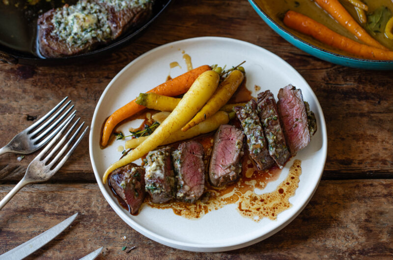 Venison haunch steaks on a white plate cooked medium-rare alongside braised carrots and green peppercorn butter.