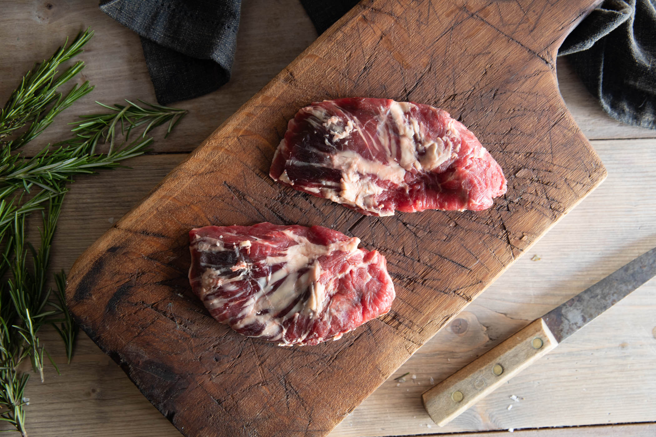 Bird's eye view of raw spider steaks on a wooden board with rosemary and a knife either side.