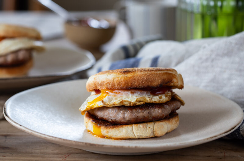 Sausage and egg muffin on off-white plate, viewed side-on. White and blue tea towel behind, ketchup and coffee mug in background.