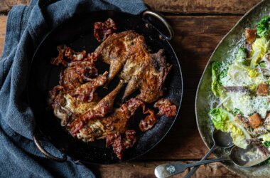 An overhead shot of a pan with two spatchcocked partridges and crispy bacon. A plate of Caesar salad to the side.