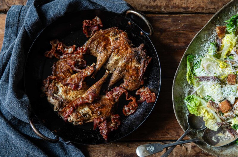 An overhead shot of a pan with two spatchcocked partridges and crispy bacon. A plate of Caesar salad to the side.