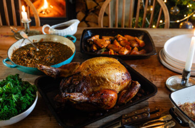 Roasted cockerel in roasting tray on farmhouse table, surrounded by accompaniments - kale to the left, roasted pumpkin, braised lentils and gravy jug behind. Lit candle and stack of off-white plates to the right, cutlery in the foreground. Wood burner with blazing fire in background, with a decorated Christmas tree beside it to the right.