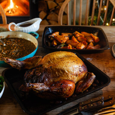 Roasted cockerel in roasting tray on farmhouse table, surrounded by accompaniments - kale to the left, roasted pumpkin, braised lentils and gravy jug behind. Lit candle and stack of off-white plates to the right, cutlery in the foreground. Wood burner with blazing fire in background, with a decorated Christmas tree beside it to the right.