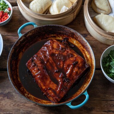 A large pan with a glazed pork belly, surrounded by steamed bao buns, pickled vegetables and picked coriander