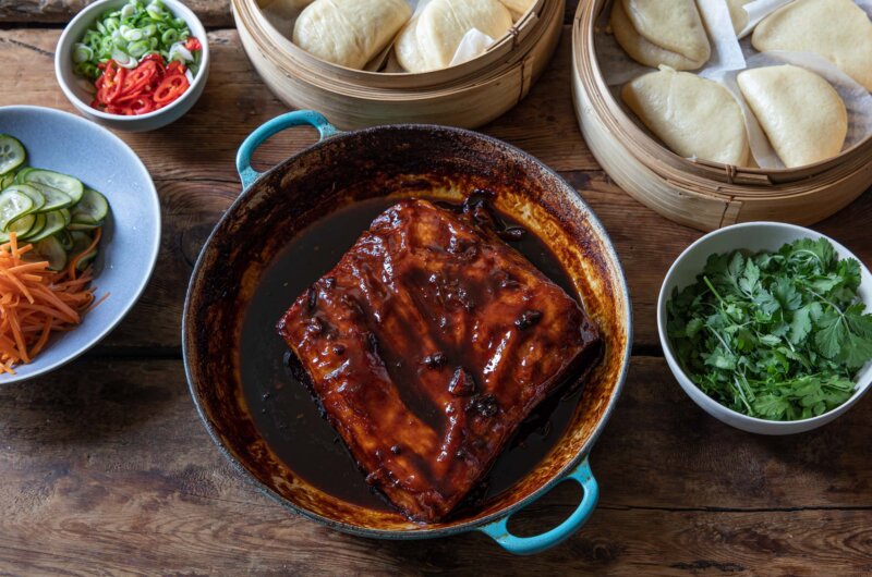A large pan with a glazed pork belly, surrounded by steamed bao buns, pickled vegetables and picked coriander