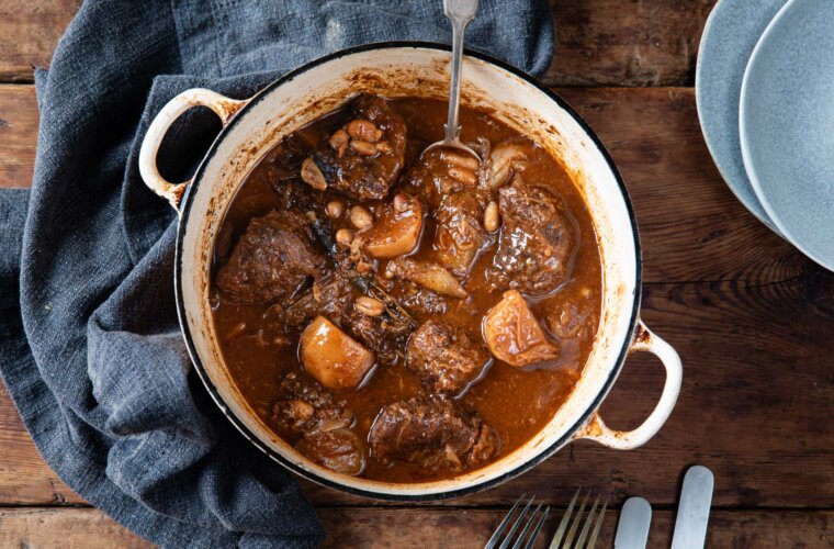 Ox cheeks, turnips and flagolet beans braised in sauce, inside a high-sided, white cast iron pan, viewed from above. Dark tea towel around half the perimeter of the pan to the left, cutlery beneath, two pale blue empty bowls top right.