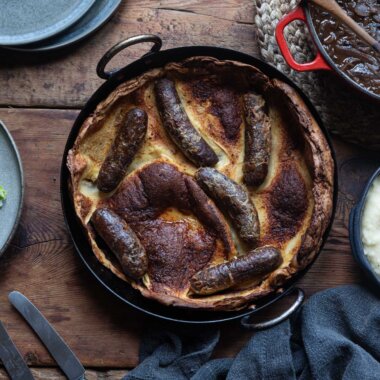 An overhead shot of a toad in the hole in a cast iron skillet, with a plate of cabbage, a bowl of mash potato and a pot of onion gravy surrounding it.