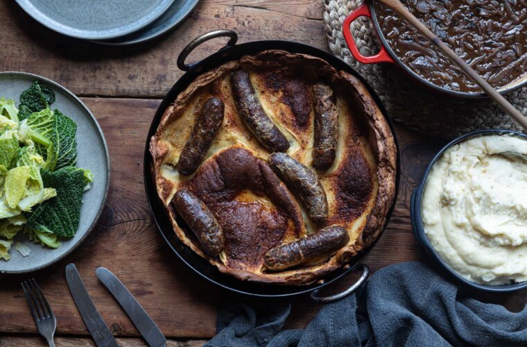 An overhead shot of a toad in the hole in a cast iron skillet, with a plate of cabbage, a bowl of mash potato and a pot of onion gravy surrounding it.