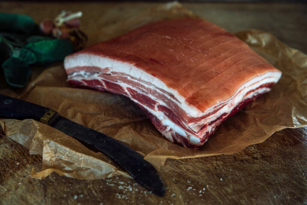 An aged pork belly on some butchers paper, on a wooden block, with a knife laid next to it. 