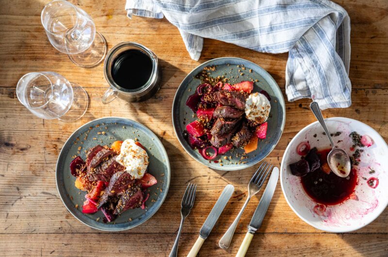 An overhead shot of a table with two plates of onglet steak, beetroots and horseradish, plus a carafe of wine and two glasses to the side.
