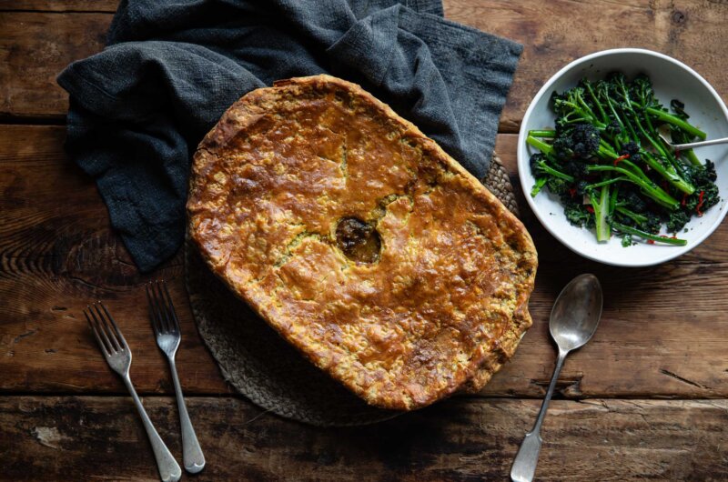 Overhead shot of a suet crusted mutton pie, with cutlery and a bowl of broccoli around.