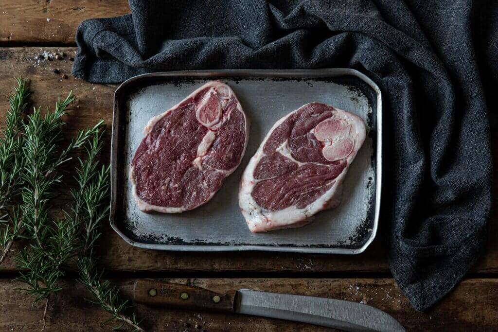 An overhead shot of two lamb ossobuco steaks, laid on a metal tray with some rosemary sprigs next to it.