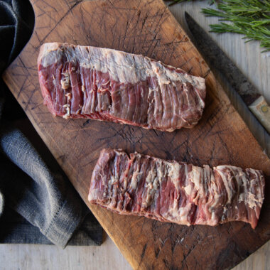Steaks viewed from above, lying on a dark, rustic chopping board. Dark tea towel to the left, vintage butcher's knife to the right. sprigs of rosemary on the extreme right.