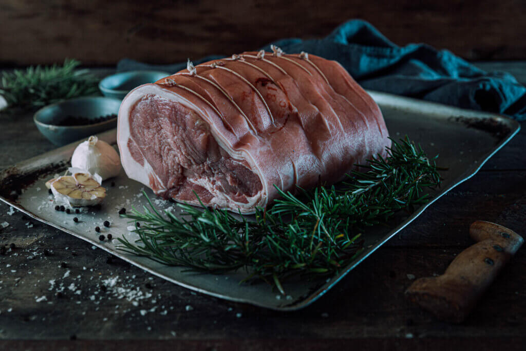 A side on photo of a rolled pork leg joint which is sat on a metal roasting tray and surrounded by sprigs of rosemary.