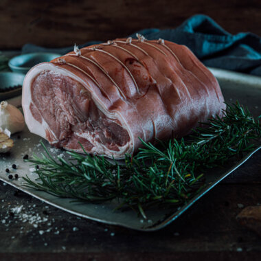 A side on photo of a rolled pork leg joint which is sat on a metal roasting tray and surrounded by sprigs of rosemary.