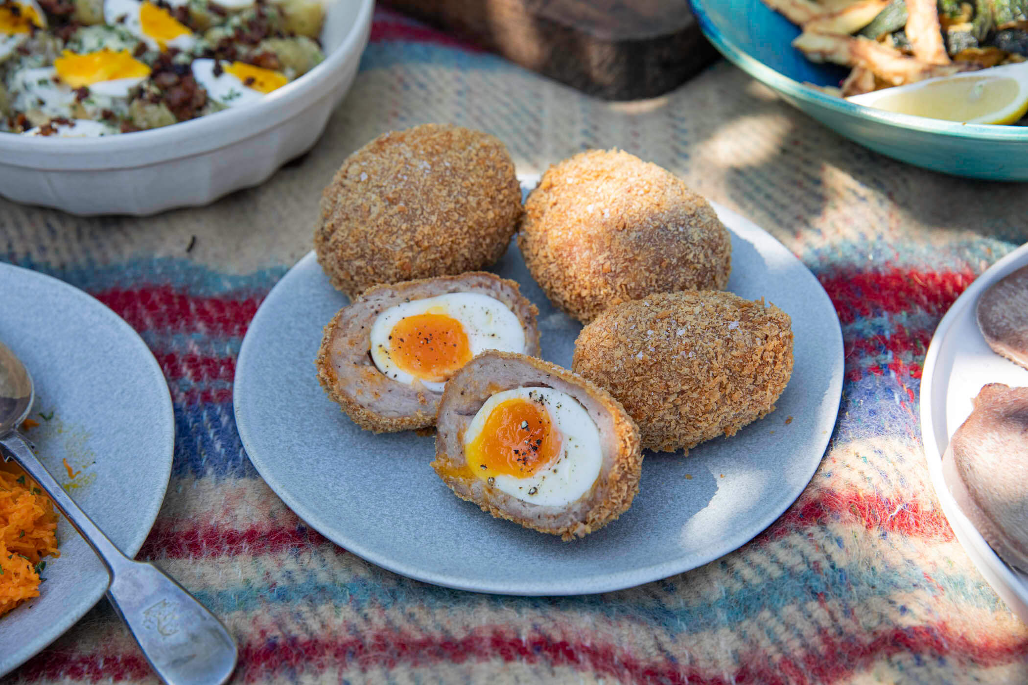 A photo of four scotch eggs on a plate, one of them is cut in half. The plate is laid on a picnic blanket, with dappled sunlight.