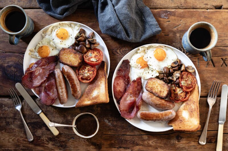 A plated Full English breakfast featuring sausages, bacon, fried eggs, tomatoes, mushrooms, fried bread, and homemade hash browns.