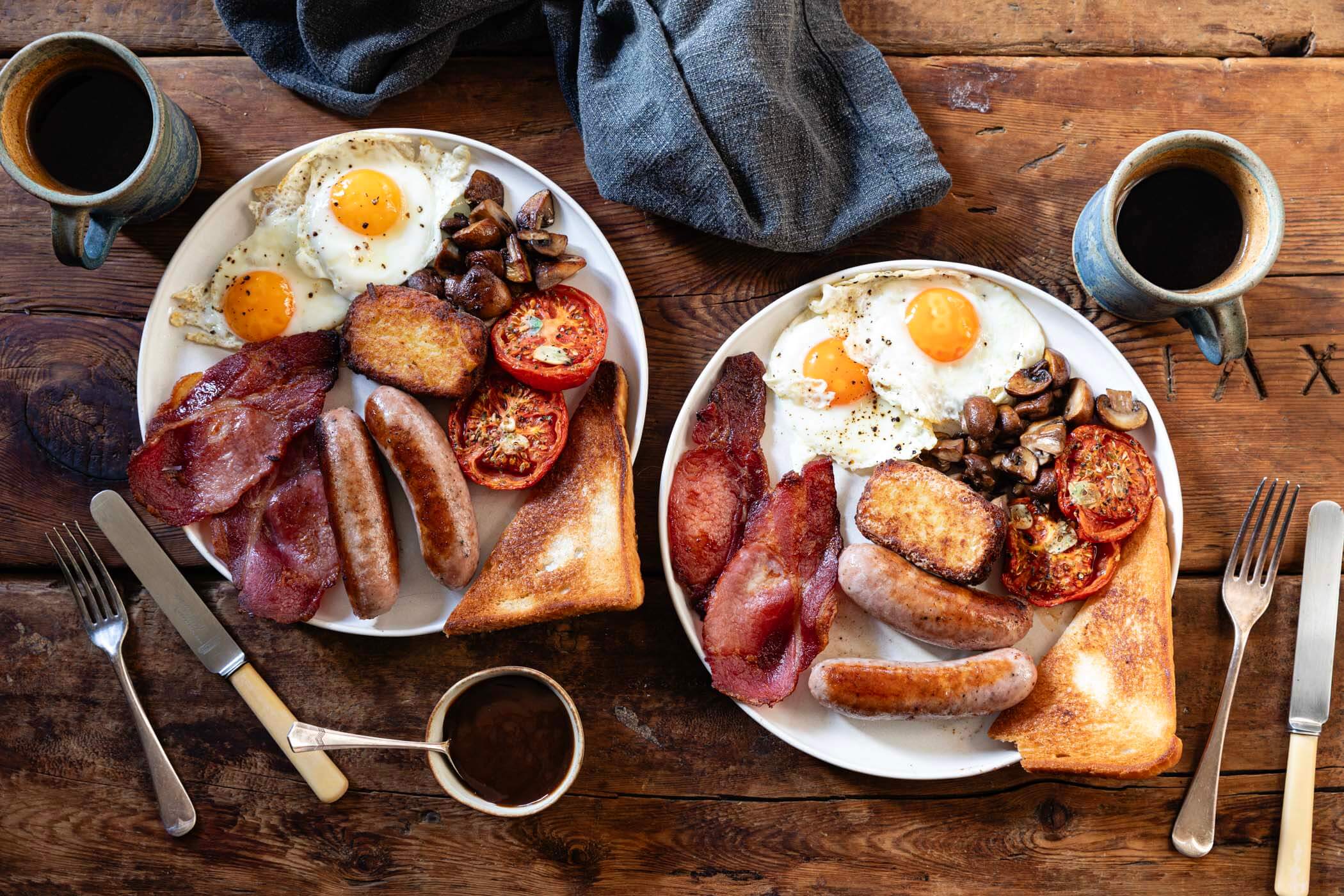 A plated Full English breakfast featuring sausages, bacon, fried eggs, tomatoes, mushrooms, fried bread, and homemade hash browns.