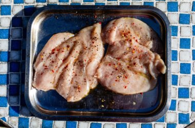 Raw butterflied chicken breast, seasoned with salt, chilli flakes and olive oil, displayed in a stainless steel cooking tray.
