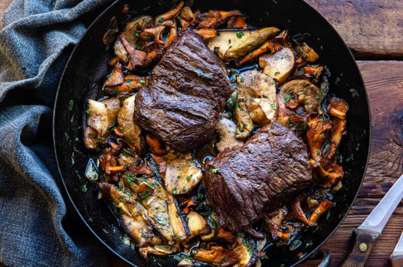 Two bavette steaks cooking in a cast iron skillet with golden ceps, garlic and parsley
