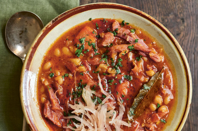 A bowl of Balkan bean čorba stew served with a spoon, showing tender beans, ham hock, and sour cabbage in a rich paprika broth