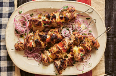 Four cooked ražnjići pork and plum skewers served on a plate with marinade, herbs and sliced onions.