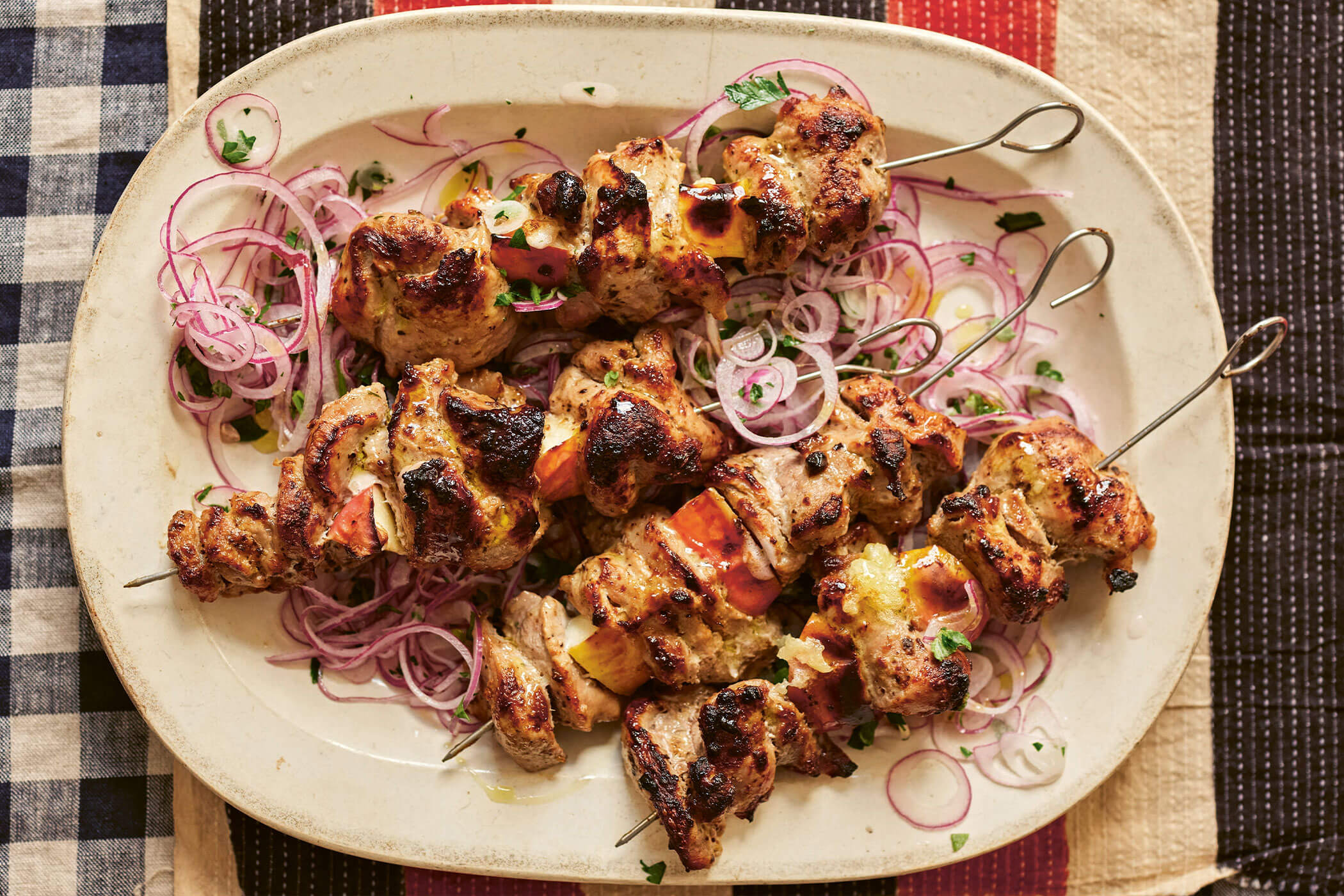 Four cooked ražnjići pork and plum skewers served on a plate with marinade, herbs and sliced onions.