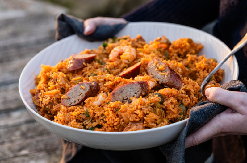 A large white bowl of Creole-style chicken and sausage jambalaya with sliced Texan-style hot links and king prawns, held in both hands