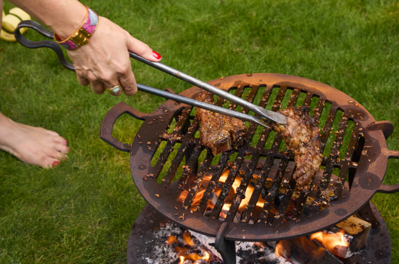 Spiced lamb chops being turned with long tongs over an open wood-fired barbecue