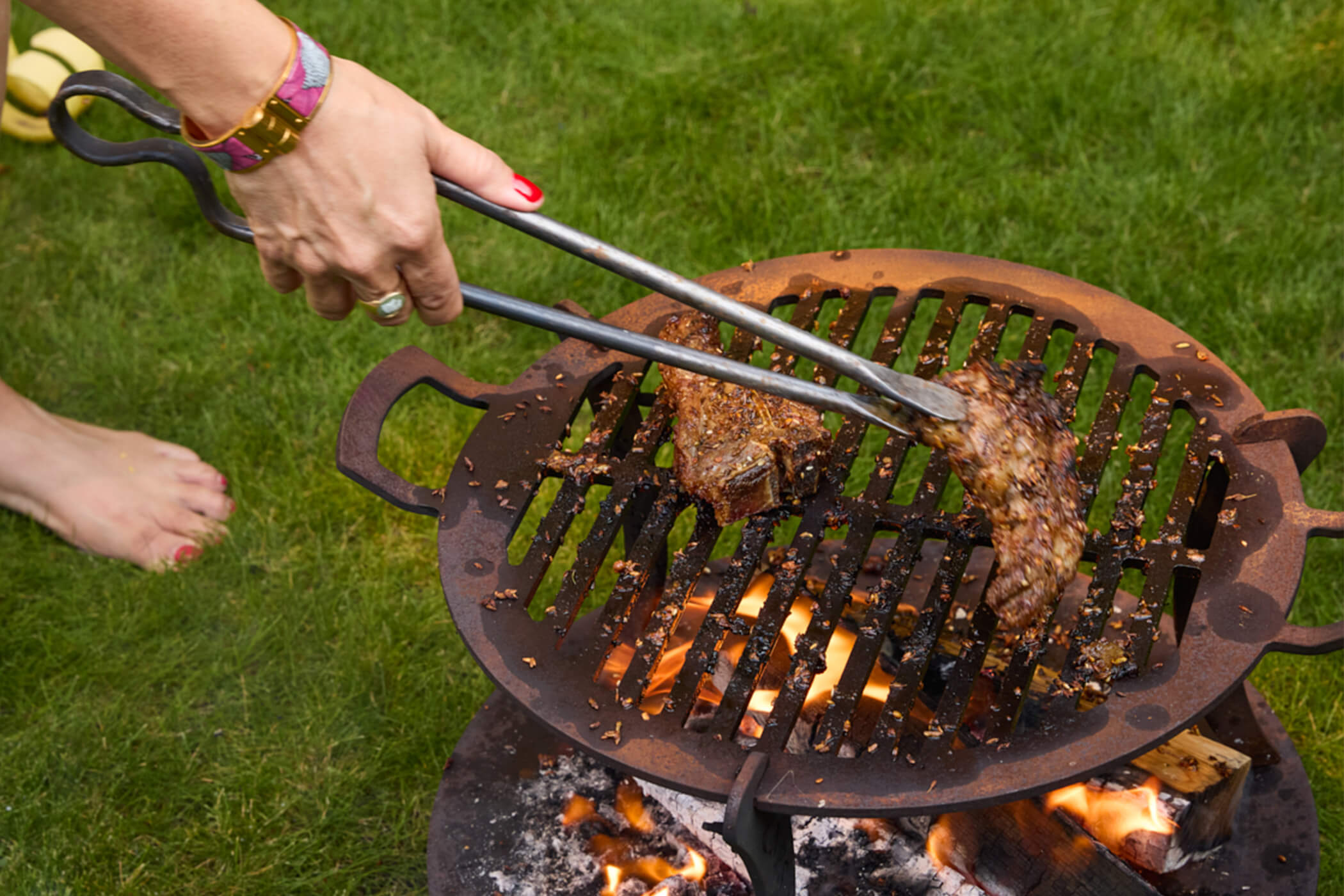 Spiced lamb chops being turned with long tongs over an open wood-fired barbecue