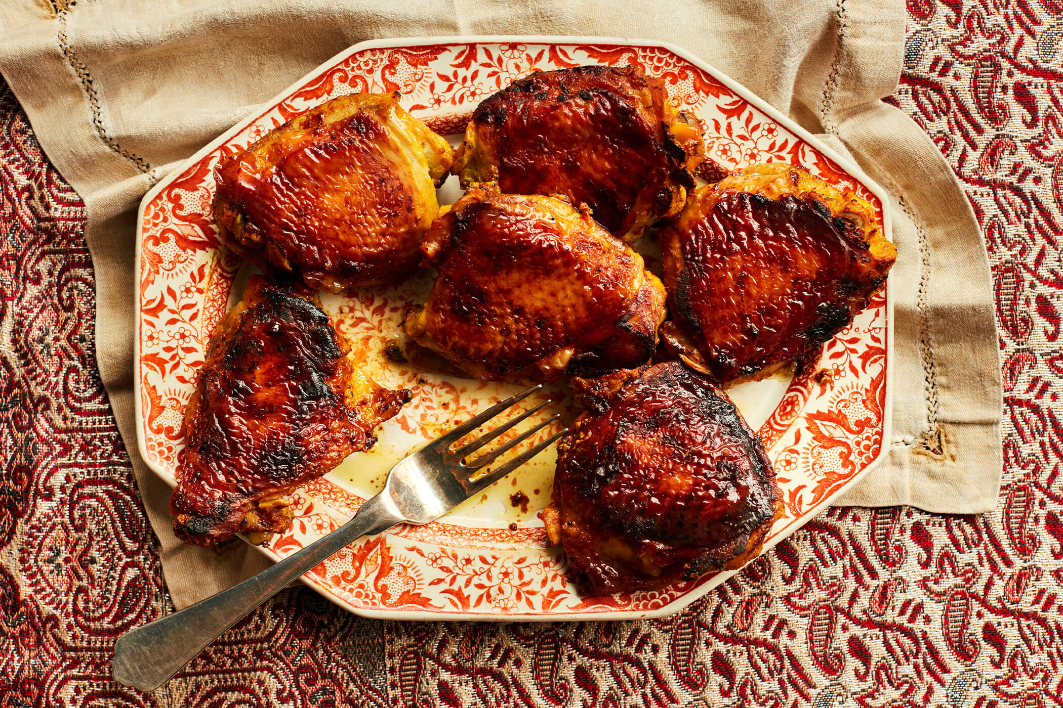 Six sticky glazed chicken thighs roasted until browned and served on a red patterned platter with a fork