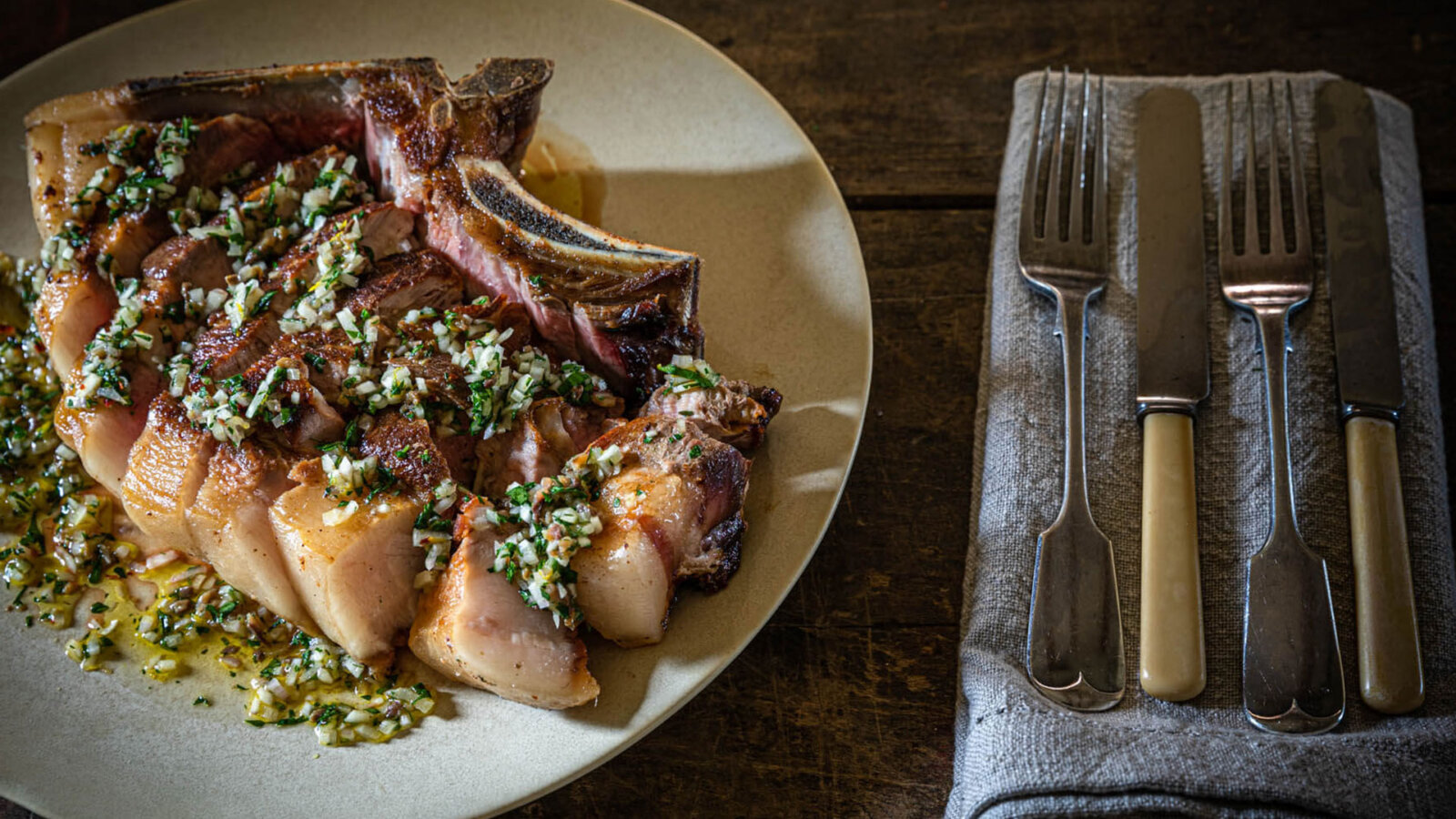 A plated pork shoulder chop with anchovy & lemon dressing with cutlery beside on a wooden table