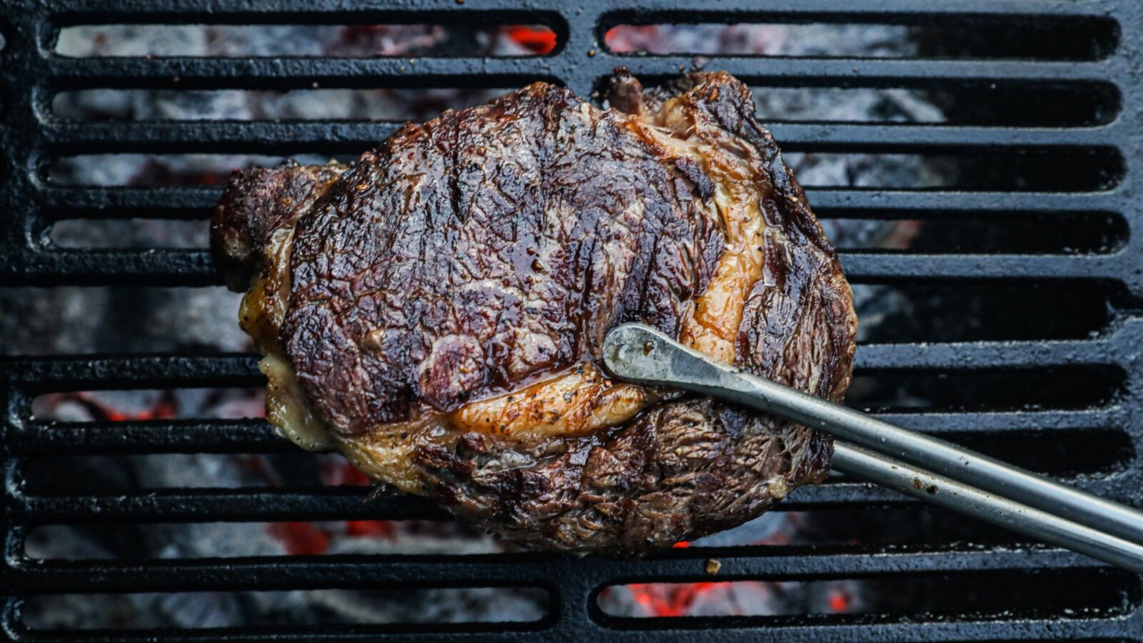A Swaledale ribeye steak held with tongs being grilled over a BBQ